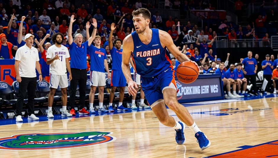 A dynamic basketball court scene featuring a 6'9" forward, Domen Petrovic, showcasing his athletic talent. In the foreground, he is dribbling a basketball, wearing a vibrant Florida Gators jersey and athletic shorts, with focused determination on his face. The middle ground includes teammates cheering him on and a coach observing intently, all in casual sports attire. In the background, the iconic Gators logo is visible on the court, with a stadium filled with excited fans. The image is bathed in warm, dramatic lighting that highlights the action and creates an engaging atmosphere, captured from a low angle to emphasize Domen's height and skill. The mood is energetic and inspiring, reflecting his journey and dedication to joining the Gator family.